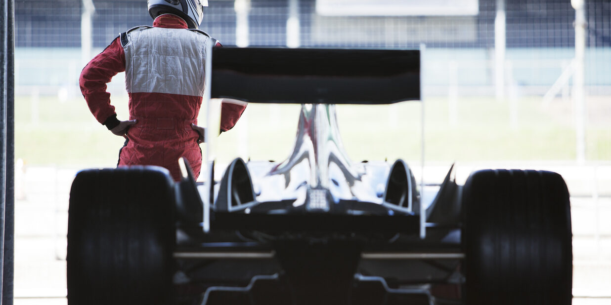 Racer standing with car in garage A race car driver stands in front of a race car parked in a garage.