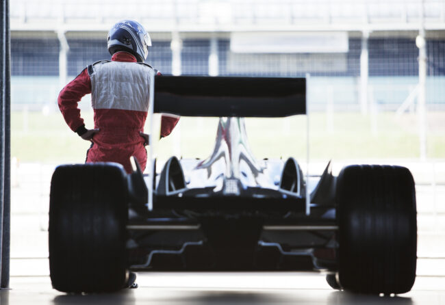 Racer standing with car in garage A race car driver stands in front of a race car parked in a garage.